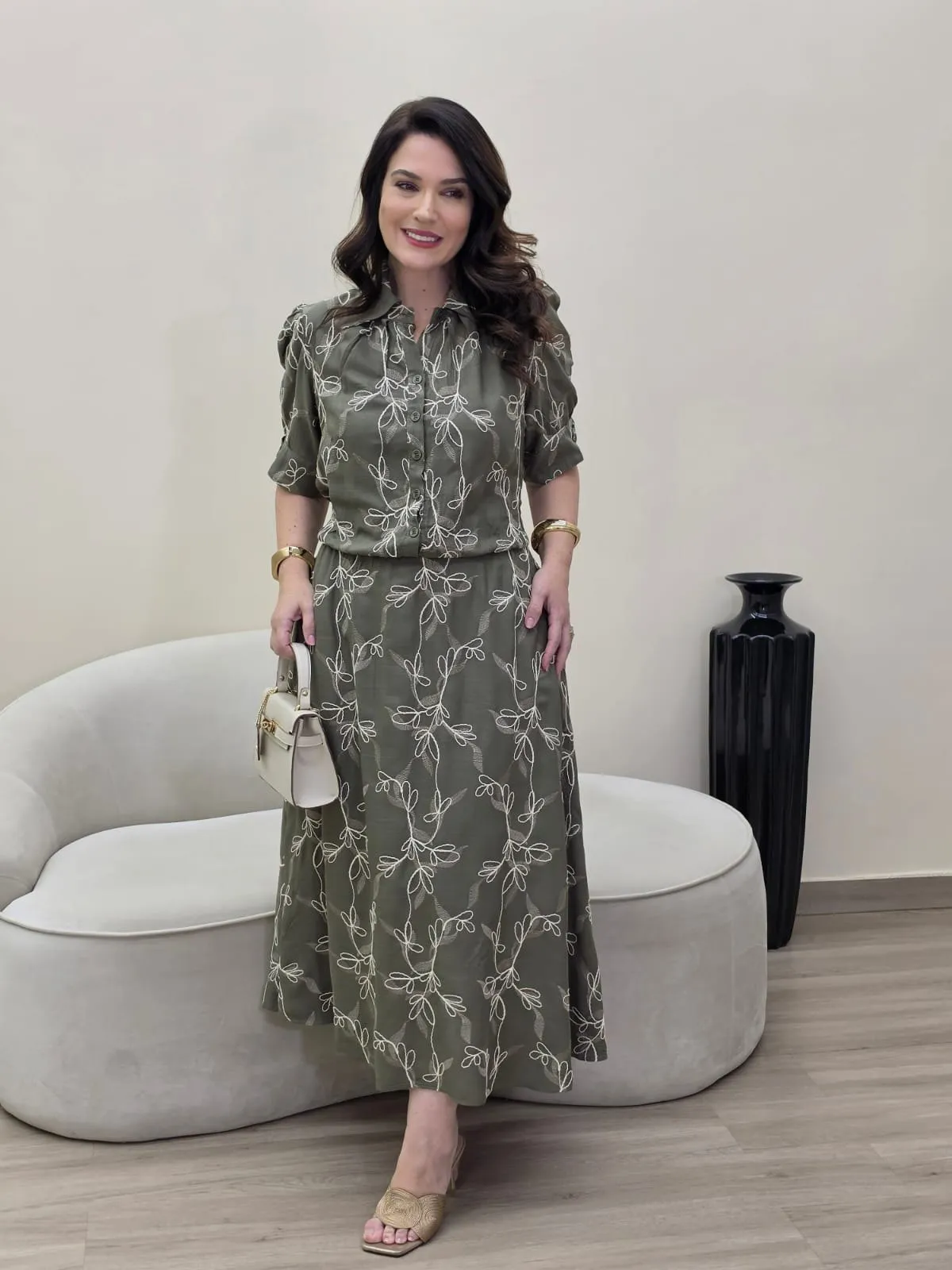 Modelo veste conjunto verde-oliva de blusa com gola e saia comprida, ambos com bordado floral branco