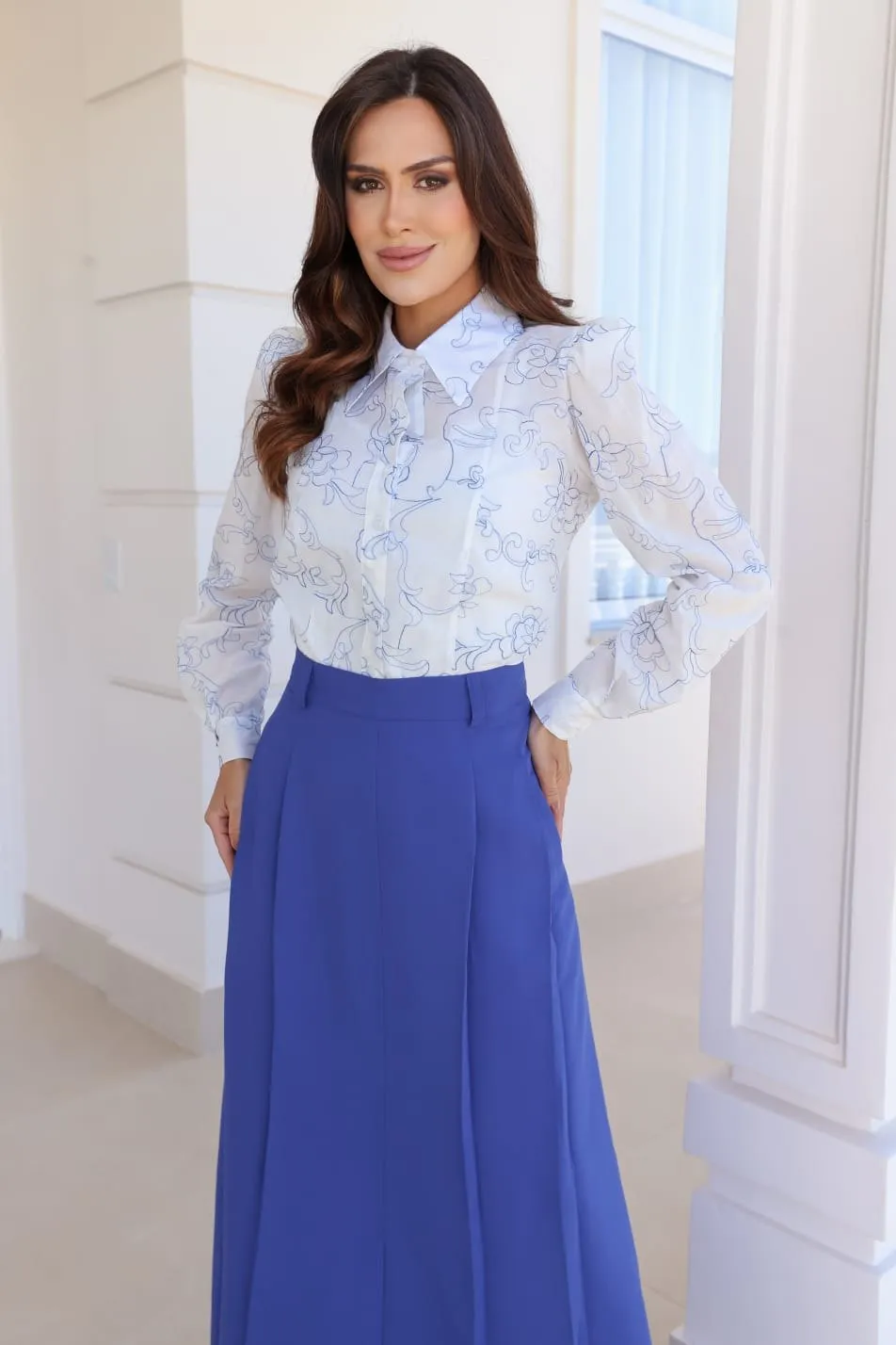 Modelo veste blusa branca em renda bordada com gola e laço, e saia comprida azul-royal