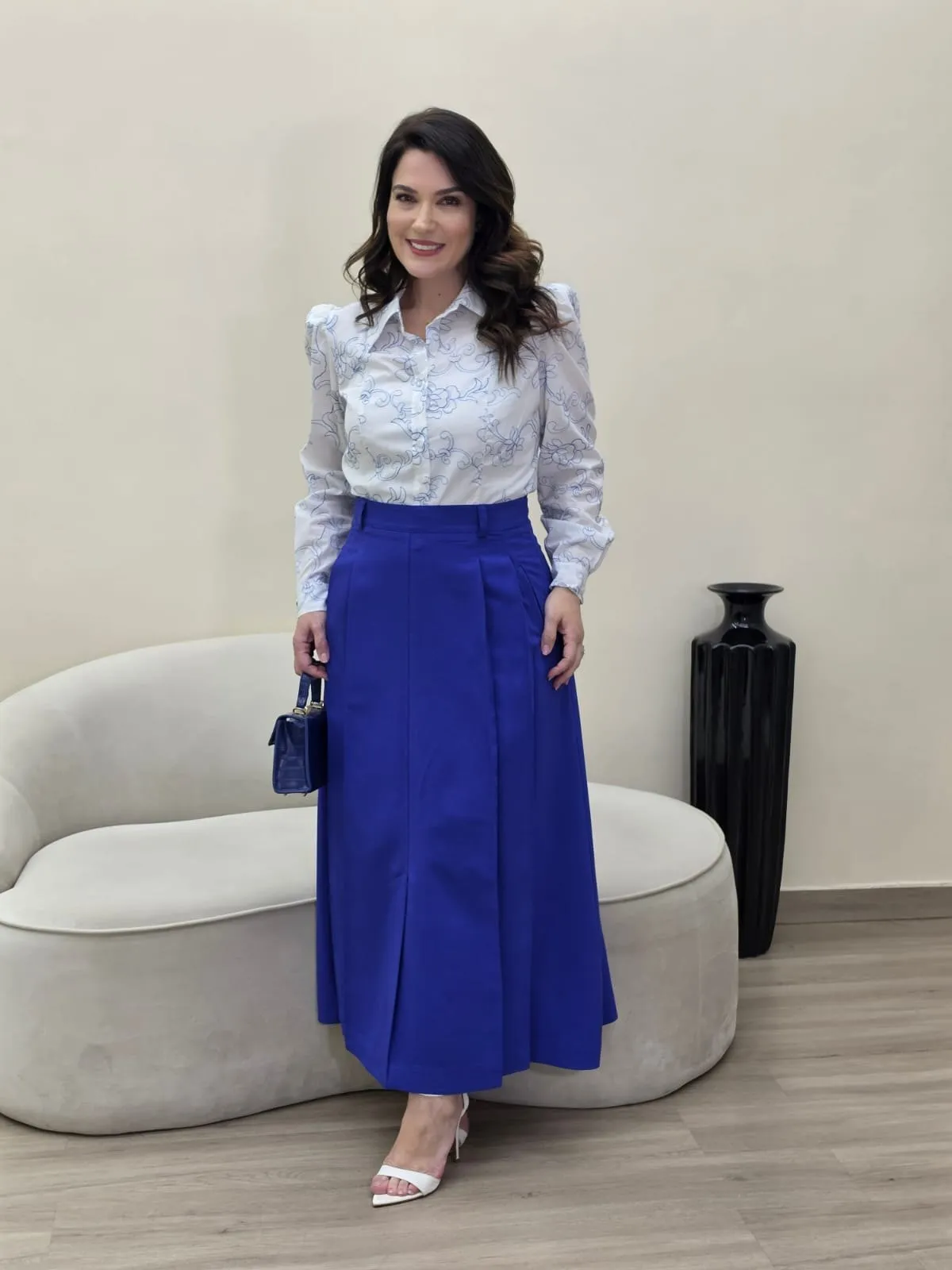 Modelo veste blusa branca em renda bordada com gola e laço, e saia comprida azul-royal