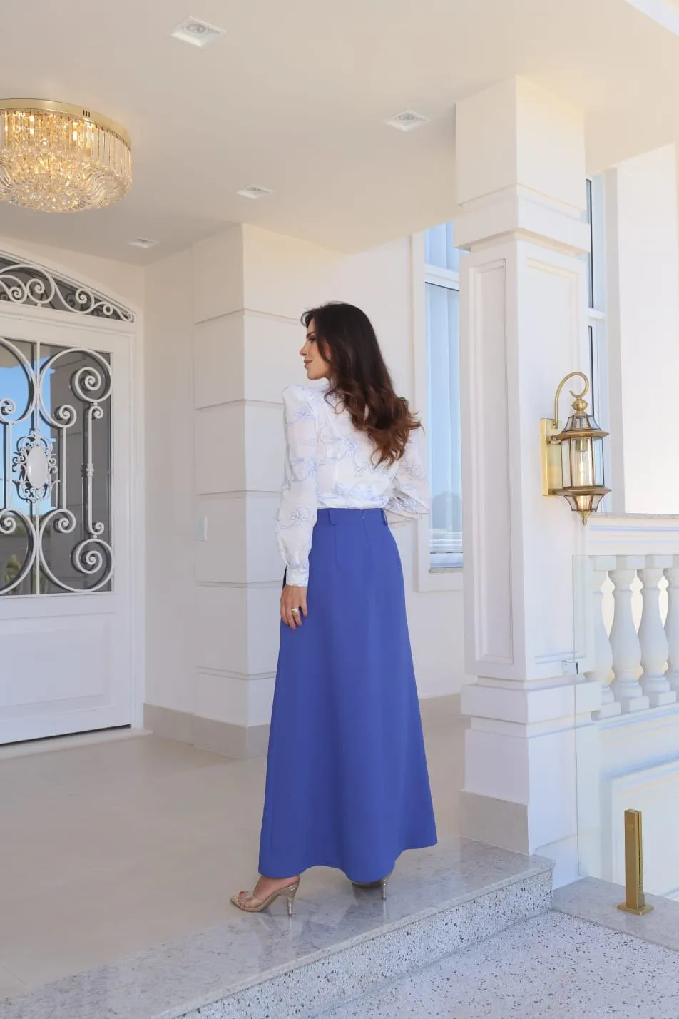 Modelo veste blusa branca em renda bordada com gola e laço, e saia comprida azul-royal